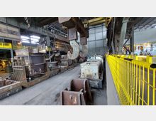 Innenansicht einer großen Industriehalle mit Schienensystemen, einem schweren Kran mit zwei massiven Metallhaken und verschiedenen Arbeitsmaschinen wie Waggons und Behältern. Gelbe Sicherheitsgeländer trennen den Arbeitsbereich von einem Gehweg, und Tageslicht fällt durch die Oberlichter und Fenster.