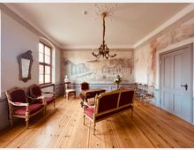 Ein historischer Salon mit Holzdielenboden, antiken Möbeln in Rot- und Goldtönen, einer verzierten Wandmalerei mit architektonischen Motiven, einem großen Fenster, einem Kronleuchter und einem dekorativen Spiegel an der Wand.