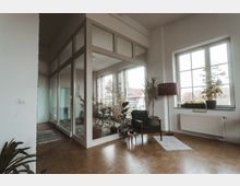 Heller Innenraum mit Holzboden, einer großen Fensterfront, einem grünen Sessel neben einer roten Stehlampe und mehreren Zimmerpflanzen. Im Hintergrund befindet sich ein abgetrennter Bereich mit Glaswänden und weiteren Pflanzen.