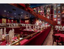 Ein eleganter Theatersaal mit dunkelroter Einrichtung, gedeckten Tischen und gefalteten Stoffservietten. Die Wände sind mit Postern und Kostümen dekoriert, eine geschwungene Holztreppe führt zu einer erhöhten Ebene.