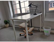 Ein einfacher Arbeitsplatz mit einem weißen Tisch, einem ergonomischen Holzstuhl und einer schwarzen Schreibtischlampe. Im Hintergrund sind große Fenster, ein Heizkörper und eine Topfpflanze sowie ein Papierkorb aus Kunststoff sichtbar.