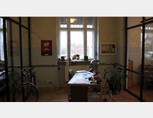 Ein Büro mit Holzboden, ausgestattet mit einem einfachen Schreibtisch, Bürostuhl, Lampe und Arbeitsmaterialien. Im Hintergrund ein großes Fenster mit Blick nach draußen sowie gläserne Trennwände auf beiden Seiten; ein Fahrrad steht links an der Wand.