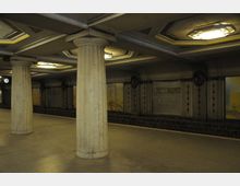 U-Bahn-Station mit Säulen im klassischen Stil, dekorativen Deckenleuchten und Wandfliesen. An der Wand ist das Stationsschild "Breitenbachplatz" zu sehen, neben Wandmalereien in gedeckten Farben.
