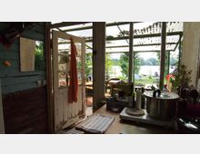 Kleines Kücheninterieur in einem rustikalen Gebäude mit Blick auf einen Garten und einen See durch große, verglaste Türen und Fenster. Auf der Arbeitsfläche stehen Küchenutensilien und ein großer Topf, die offene Tür führt zu einem Außenbereich mit Tischen und Bäumen.