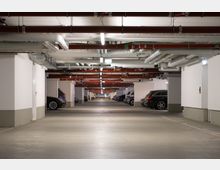Unterirdische Parkgarage mit weißen Wänden und freiliegenden roten und weißen Rohrleitungen an der Decke. Auf beiden Seiten sind geparkte Autos, und der Boden besteht aus glattem Beton.