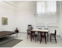 Ein minimalistischer Raum mit weißen Backsteinwänden und hohen, lichtdurchfluteten Fenstern mit weißen Vorhängen. Im Vordergrund stehen dunkle Holzstühle und Tische, daneben eine kleine Bühne; an der Wand hängt ein Gemälde, und in der Ecke befindet sich ein Lautsprechersystem.