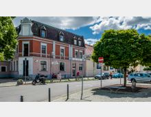 Straßenszene mit einem Eckgebäude aus mehrstöckigem rotem Backstein mit weißen Verzierungen und einem dunklen Dach aus Schindeln, darunter Schilder mit Straßennamen. Im Vordergrund stehen ein Baum, ein Motorrad und geparkte Autos; daneben fährt eine Person auf einem Fahrrad.