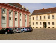Historischer Innenhof mit gepflastertem Boden, umgeben von zwei Gebäuden im klassischen Architekturstil. Links ein rosa und cremefarbenes Gebäude mit Säulen und Statuen auf dem Dach, rechts ein gelbes Gebäude mit verzierten Fensterrahmen; geparkte Autos im Vordergrund.