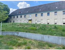Zweistöckiges ehemaliges Gebäude mit grauer Putzfassade und einfachem Satteldach, Fenster teils vergittert oder zugemauert. Im Vordergrund eine leicht abschüssige, grasbewachsene Fläche mit einem niedrigen Betonsockel; links ein einzelner Baum.