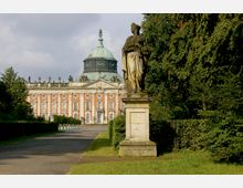 Ansicht des Neuen Palais im Park Sanssouci in Potsdam, mit einer barocken Skulptur im Vordergrund, flankiert von symmetrischen Hecken und Bäumen. Das Gebäude zeigt eine Fassade mit rosa und weißen Elementen sowie eine grüne Kuppel.
