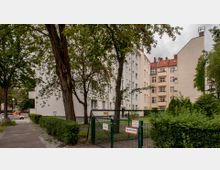 Mehrstöckige Wohngebäude mit heller Fassade, umgeben von Bäumen und Grünflächen. Im Vordergrund ein Metallzaun mit einem Tor, das mit einem Schild „Feuerwehrzufahrt“ gekennzeichnet ist, sowie ein Gehweg entlang der linken Seite.