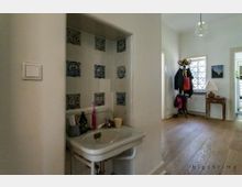 Innenraum eines Hauses mit einem kleinen Waschbecken an der Wand links, umgeben von weiß-blauen Fliesen. Im Hintergrund ein Flur mit Holzfußboden, einer Garderobe mit Jacken und einem kleinen Tisch mit Lampe vor einem Fenster mit Gitter.