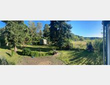 Gartenlandschaft mit großer Rasenfläche, Bäumen und Büschen, im Hintergrund ein kleiner grüner Bauwagen. Rechts ist ein gemauerter Grill, im Vordergrund eine gepflasterte Terrasse mit Blumenbeeten. Dahinter erstreckt sich eine Wiese bis zum Waldrand.