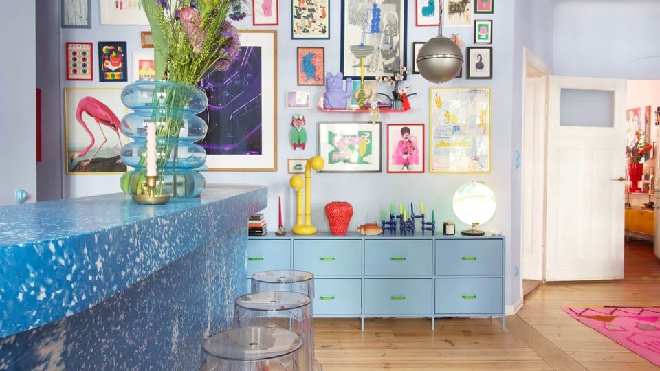 Bright, colorful interior with a speckled blue bar counter and clear stools, facing a pale blue wall covered in framed artwork above a low dresser with decorative objects and a globe lamp. A metallic pendant light hangs from the ceiling, and a doorway opens to an adjacent room with wood floors and a pink rug.