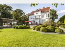 Großer Garten mit gepflegtem Rasen und runden Buchsbaumsträuchern vor einer weißen, mehrstöckigen Villa mit roten Dachziegeln. Links ist ein modernes, verglastes Gebäude mit Terrasse zu sehen, im Hintergrund befinden sich Bäume und blauer Himmel.