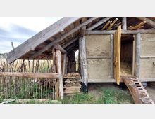 Holzhaus mit Strohdach in traditioneller Bauweise, bestehend aus Holzbalken, Lehmwänden und einem offenen Verschlag zur Lagerung von Holz. Im Vordergrund ein Lattenzaun aus Ästen und eine schmale Holzbrücke, die zu einer halb geöffneten Tür führt.