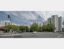 Straßenkreuzung mit einer Tankstelle links im Bild und mehreren Hochhäusern im Hintergrund, die von Bäumen umgeben sind; Ampeln und parkende sowie fahrende Autos prägen die Szene. Der Himmel ist leicht bewölkt.