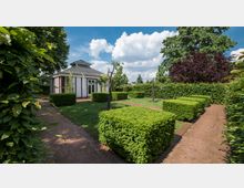 Ein gepflegter Garten mit symmetrisch geschnittenen Buchsbaumhecken, Rasenflächen und schmalen Gehwegen aus rotem Kies. Im Hintergrund steht ein kleines Gebäude mit einem flachen Dach und Glasfronten, umgeben von hohen Bäumen und Hecken.