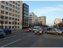 Eine Stadtstraße mit mehrspurigem Verkehr, flankiert von modernen Bürogebäuden mit großen Fenstern. Im Vordergrund fahren Autos und ein Kleintransporter, während kahle Bäume und Straßenlaternen am Gehweg stehen.