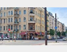 Straßenszene mit einem vierstöckigen Altbau mit beige-gelber Fassade und runden Fenstern. Im Erdgeschoss befindet sich ein Restaurant mit dem Namen „Antalya Bistro“, erkennbar durch rote Markisen und eine Außenbestuhlung; im Vordergrund eine Kreuzung mit Verkehrszeichen und geparkten Autos.