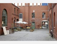 Gepflasterter Innenhof eines historischen Backsteingebäudes mit großen Fenstern und einem grünen Tor, beschriftet mit „Klempner“. Im Hof stehen Schirme, Pflanzkästen, Fahrradständer und Schilder, darunter eines mit der Aufschrift „Alte Kantine“.