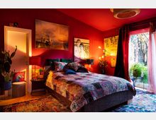 Ein Schlafzimmer mit roten Wänden und warmem Licht, ausgestattet mit einem großen, gemusterten Bett und dekorativen Kissen. An den Wänden hängen Gemälde, eine Glastür auf der rechten Seite führt zu einem Außenbereich mit Pflanzen.