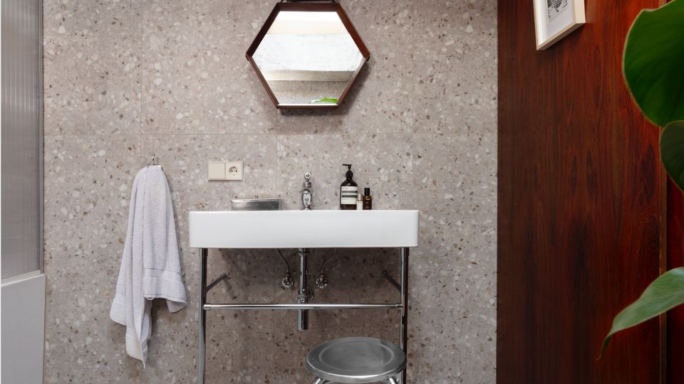 Modern bathroom vanity area with terrazzo walls and floor, featuring a white rectangular sink on chrome legs, a hexagonal mirror, and a towel hanging beside the basin. A dark wood-paneled wall with a framed picture contrasts with the concrete ceiling and a small metal stool in front of the sink.