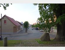 Straßenszene in einer kleinen Stadt mit gepflasterter Fahrbahn, links und rechts von niedrigen Wohnhäusern mit roten Ziegeldächern flankiert. Im Vordergrund eine Grünfläche mit einem großen Baum und einem Findling.