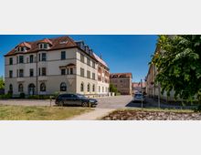 Straßenszene in einem Wohnviertel mit einem mehrstöckigen Altbau mit hellem Putz, Erkern und rotem Ziegeldach an einer Ecke. Rechts verläuft eine ruhige Straße mit weiteren Gebäuden, parkenden Autos und einem Baum am Gehweg unter blauem Himmel.