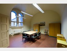 Ein schlichter Besprechungsraum mit gewölbter Decke und zwei großen Fenstern mit Blick auf ein benachbartes Gebäude. Der Raum enthält einen rechteckigen Tisch mit sechs Stühlen, zwei Holzschränke, eine Heizung unter den Fenstern und eine Wanddekoration in Form eines Gemäldes.