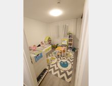 Kinderzimmer mit hellem Holzboden und einem Teppich mit Eulenmotiv. Zu sehen sind ein Kinderbett, ein kleiner Tisch mit Stühlen, ein Spielzeugregal, eine Miniaturküche und Spielzeug, sowie ein Keyboard am Boden.