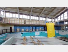 Ehemaliges Schwimmbad mit leerem Becken, abgeblätterter Wandfarbe und einer Galerie mit Umkleideschränken. Ein großes gelbes Tuch hängt von der Galerie, und im Beckenbereich stehen Stühle aufgestellt.