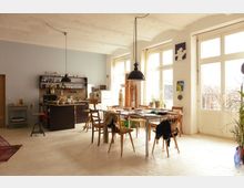 Ein heller Wohnraum mit offener Küche, Esstisch und großen Fenstern. Der Raum verfügt über helle Wände, hölzerne Möbel und Hängeleuchten, mit Zugang zu einem Balkon oder Außenbereich durch Glastüren.