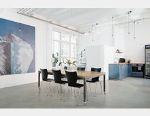 Heller, moderner Gemeinschaftsraum mit einem großen Holztisch und schwarzen Stühlen, einer Küchenzeile mit blauen Schränken und einem Getränkekühlschrank. Im Hintergrund hängen große Fenster und ein abstraktes Gemälde an weißen Wänden.