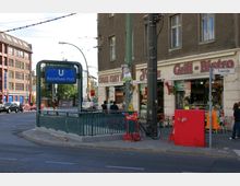 U-Bahneingang am Rosenthaler Platz in Berlin, umgeben von mehreren Imbissen, darunter ein "Grill-Bistro" und ein asiatisches Schnellrestaurant. Eine belebte Straßenecke mit Gehweg, Laternenmast und Radständer im Vordergrund.
