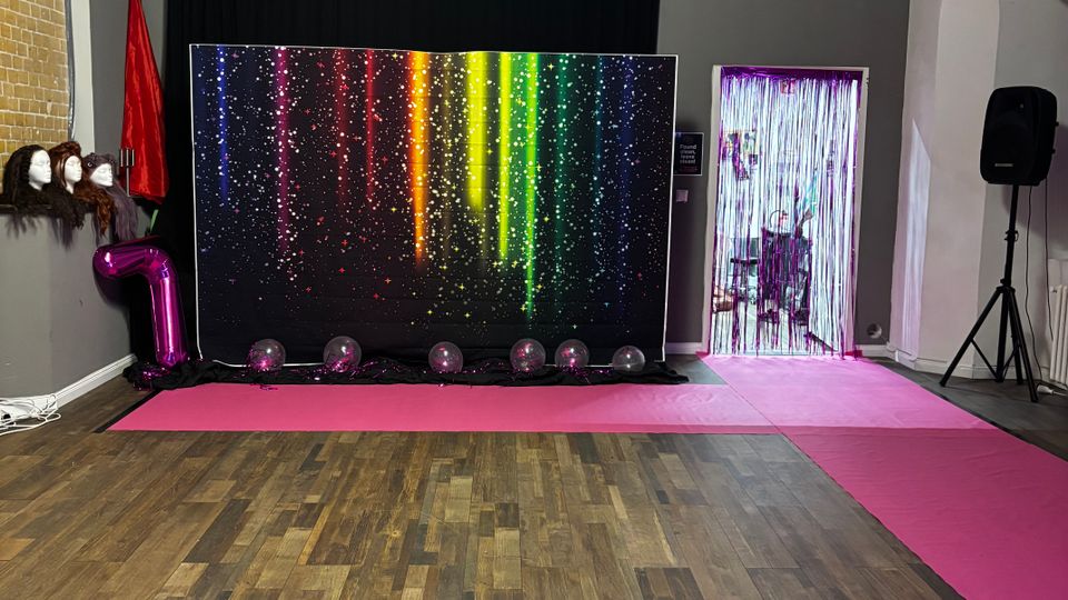 Open studio or event space with a wood plank floor and a pink carpet runway leading to a backdrop with rainbow light streak graphics in front of black curtains. The ceiling has exposed pipes and acoustic panels, and the stage area includes balloons, a large “7” balloon, a tinsel photo curtain, and a speaker on a stand.
