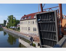 Eine angehobene Klappbrücke über einem Wasserkanal, daneben ein zweistöckiges Gebäude mit rotem Ziegeldach und großen Fenstern. Im Hintergrund sind ein Weg mit Laternen, Bäume und Sitzmöglichkeiten mit Sonnenschirmen zu sehen.