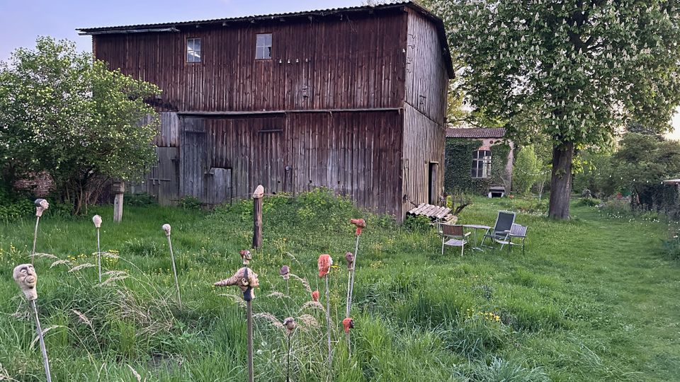 Große, verwitterte Holzscheune mit Wellblechdach in einem ländlichen Garten mit hohem Gras und blühenden Bäumen. Im Vordergrund stehen mehrere Stäbe mit kleinen Skulpturen, rechts neben einem großen Baum sind Gartenstühle auf der Wiese aufgestellt.