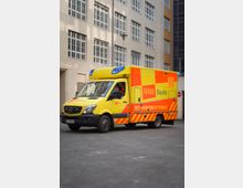 Ein gelbes Rettungsfahrzeug mit der Aufschrift "NAW Berlin" und "Rettungsdienstschule" steht auf einem Parkplatz vor einem mehrstöckigen Gebäude mit großen Fenstern. Das Fahrzeug trägt rote und orangefarbene reflektierende Markierungen.