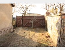 Ein kleiner, unbefestigter Hof mit einer alten, hölzernen Doppeltorstruktur im Hintergrund sowie einer Backsteinmauer rechts; links ist eine verputzte Hauswand zu sehen. Der Boden ist teilweise mit Gras und Laub bedeckt, und im Hintergrund stehen kahle Bäume.