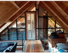 Ein Dachgeschosszimmer mit sichtbaren Holzsparren, einer Glastür mit schwarzem Metallrahmen und großen Glasfenstern, die in andere Bereiche des Hauses blicken. Der Raum ist mit Holzfußboden ausgestattet, und es sind Möbel sowie Wäschekörbe zu sehen.