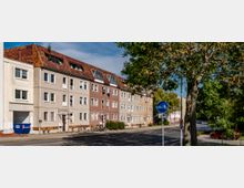 Mehrfamilienhäuser in einer Wohnstraße mit vier Etagen und Schrägdächern, teilweise mit Solarpaneelen. Im Vordergrund befinden sich ein Geh- und Radweg-Schild sowie Bäume, die die Straße säumen.