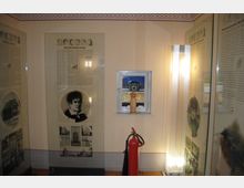 Ein Innenraum mit Ausstellungstafeln in Glasvitrinen, die beschriftete Texte und historische Fotografien zeigen. Ein kleines Fenster mit Blick nach draußen und ein roter Feuerlöscher sind ebenfalls sichtbar.