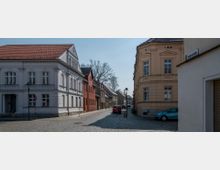Straßenszene in einer Altstadt mit gepflasterter Straße, flankiert von historischen Gebäuden mit verzierten Fassaden; links ein weißes Haus, rechts ein beiges Eckgebäude, dahinter Reihenhäuser und parkende Autos.