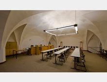 Ein historischer Raum mit Gewölbedecke, eingerichtet als Seminarraum mit langen Tischen und schwarzen Stühlen. Im Hintergrund stehen ein Rednerpult, Holzschränke und eine Lampe; die Beleuchtung besteht aus modernen rechteckigen Hängelampen.