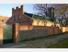 Backsteingebäude mit Satteldach und hoher, hölzerner Glockenturm im Hintergrund, umgeben von einer roten Backsteinmauer mit einem grünen Holztor. Im Vordergrund ein gepflasterter Weg und Bäume ohne Laub.