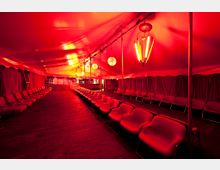 Ein Innenraum eines roten Zeltes mit Sitzreihen aus roten Stühlen, die entlang der Seiten verlaufen. Die Beleuchtung besteht aus dekorativen Hängelampen und kugelförmigen Leuchten, die ein warmes, rotes Licht erzeugen, während der Boden aus dunklem Holz besteht.