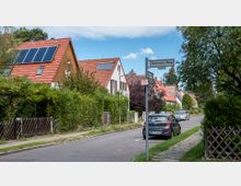 Eine ruhige Wohnstraße mit Einfamilienhäusern, die rote Ziegeldächer und teils Solarpaneele aufweisen. Die Straße ist von Bäumen und Hecken gesäumt, vereinzelt stehen Autos am Straßenrand, und ein Straßenschild mit der Bezeichnung „Smyrnaer Weg“ ist sichtbar.