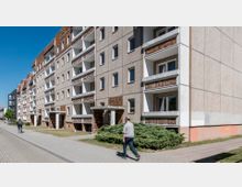 Wohnblock mit mehreren Stockwerken in typischer Plattenbauweise, bestehend aus grauem Beton mit braunen Keramikelementen an Balkonen und Eingängen. Im Vordergrund ein Fußgängerweg mit zwei Personen, einer zu Fuß und der andere auf einem Fahrrad, sowie ein kleiner Grünstreifen mit Büschen.