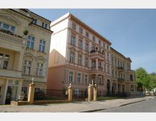 Mehrstöckiges, historisches Wohngebäude mit verzierten Fassaden und Balkonen in hellen Pastellfarben, umgeben von einem schmiedeeisernen Zaun mit gemauerten Torpfosten an einer gepflasterten Straße. Im Hintergrund sind Bäume und ein klarer Himmel zu sehen.